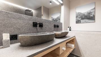 Een moderne badkamer met twee ruime stenen wastafels, een houten wastafelmeubel, een grote spiegel en een schilderij van een berglandschap aan de muur.