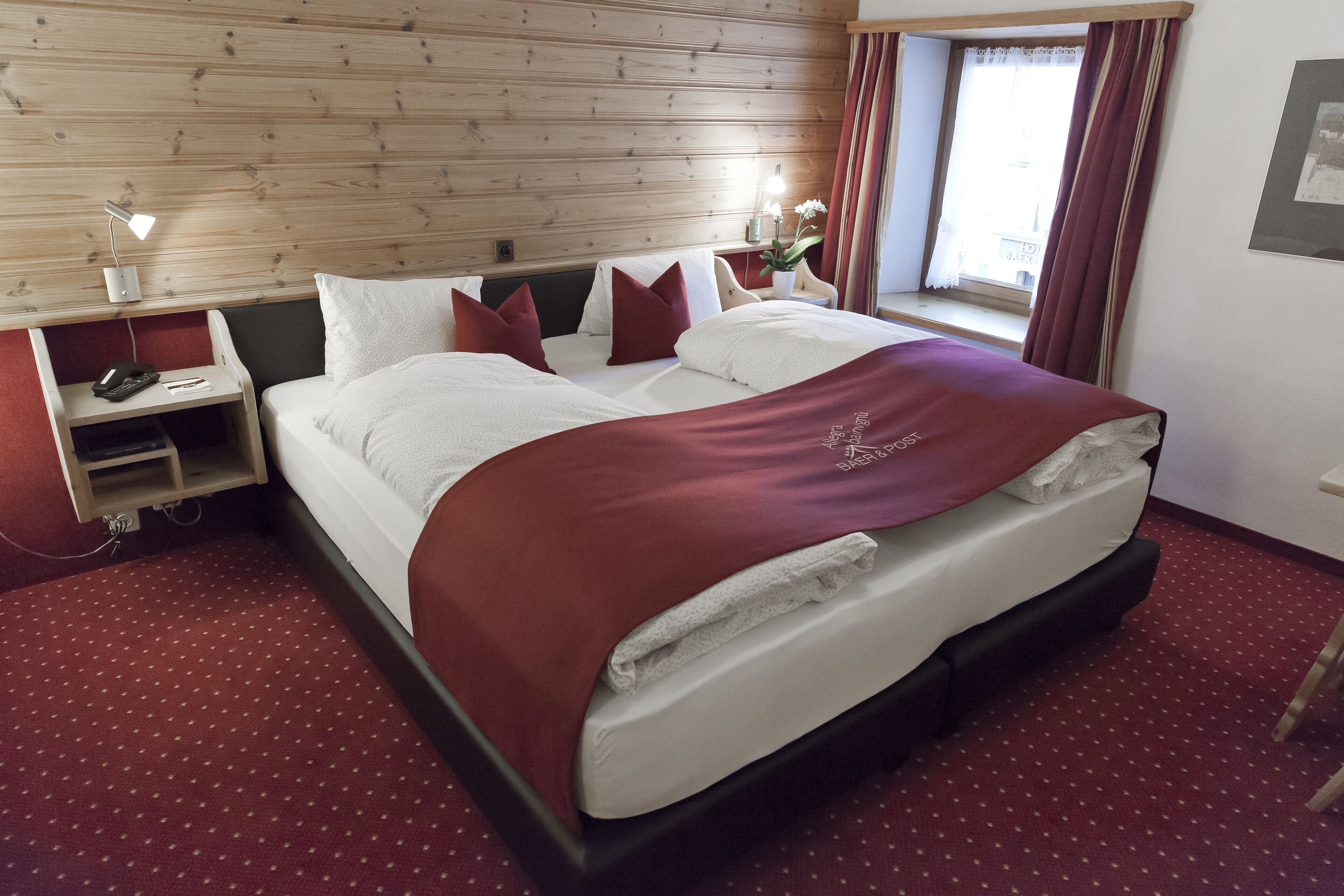 A hotel room with a wooden headboard, a large bed with white and red bedding, a nightstand with a lamp, and a window with curtains.