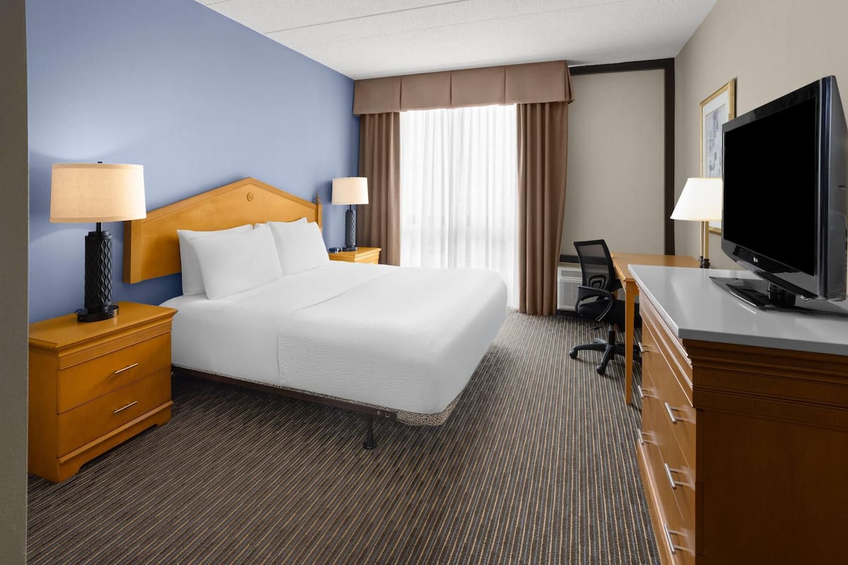 A hotel room with a bed, two bedside tables with lamps, a desk with a chair, a flat-screen TV, and a large window with curtains.