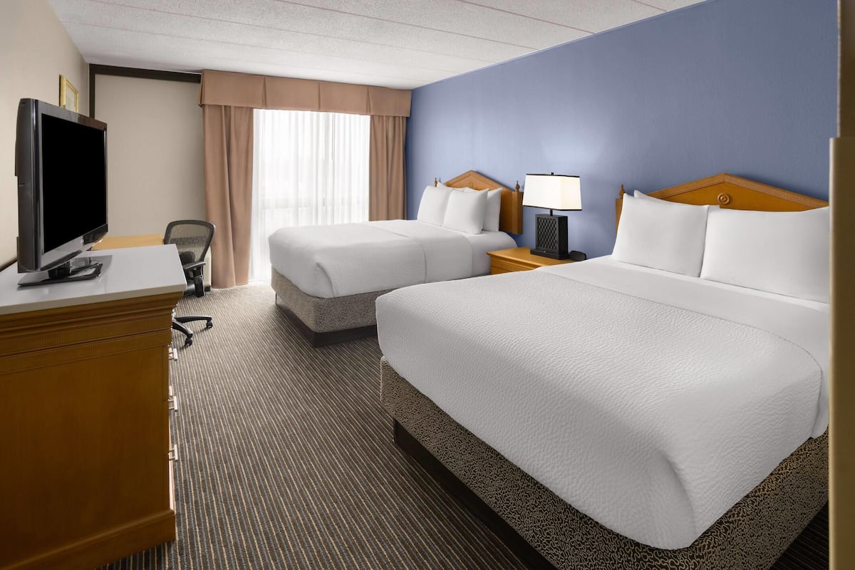 A hotel room with two beds, a desk with a TV, a chair, and a nightstand with a lamp.