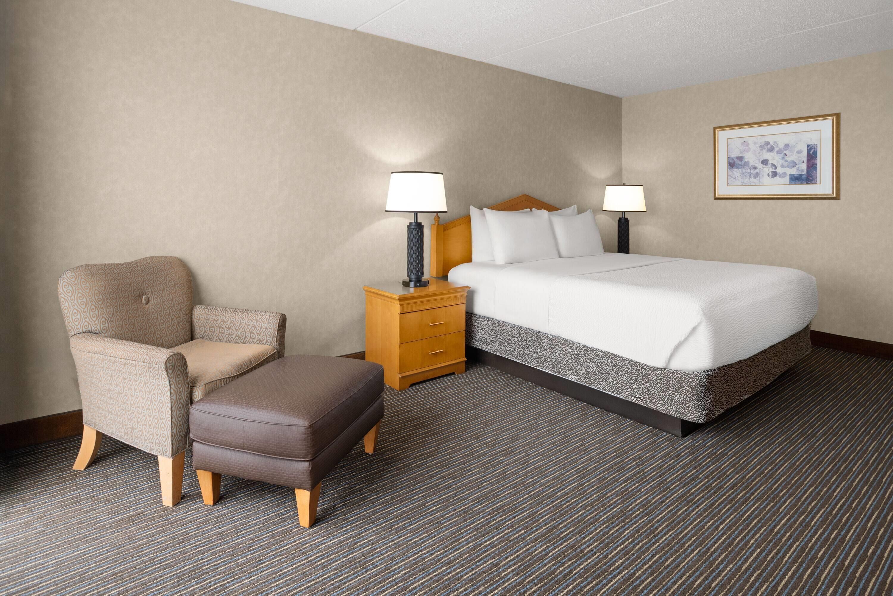 A hotel room with a bed, a chair, an ottoman, a nightstand with lamps, and a framed picture on the wall.