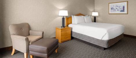 A hotel room with a bed, a chair, an ottoman, a nightstand with lamps, and a framed picture on the wall.