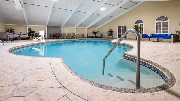 An indoor swimming pool with a tiled edge, a metal ladder, and a seating area with a sofa and chairs. - Days Hotel by Wyndham Toms River Jersey Shore (Toms River)