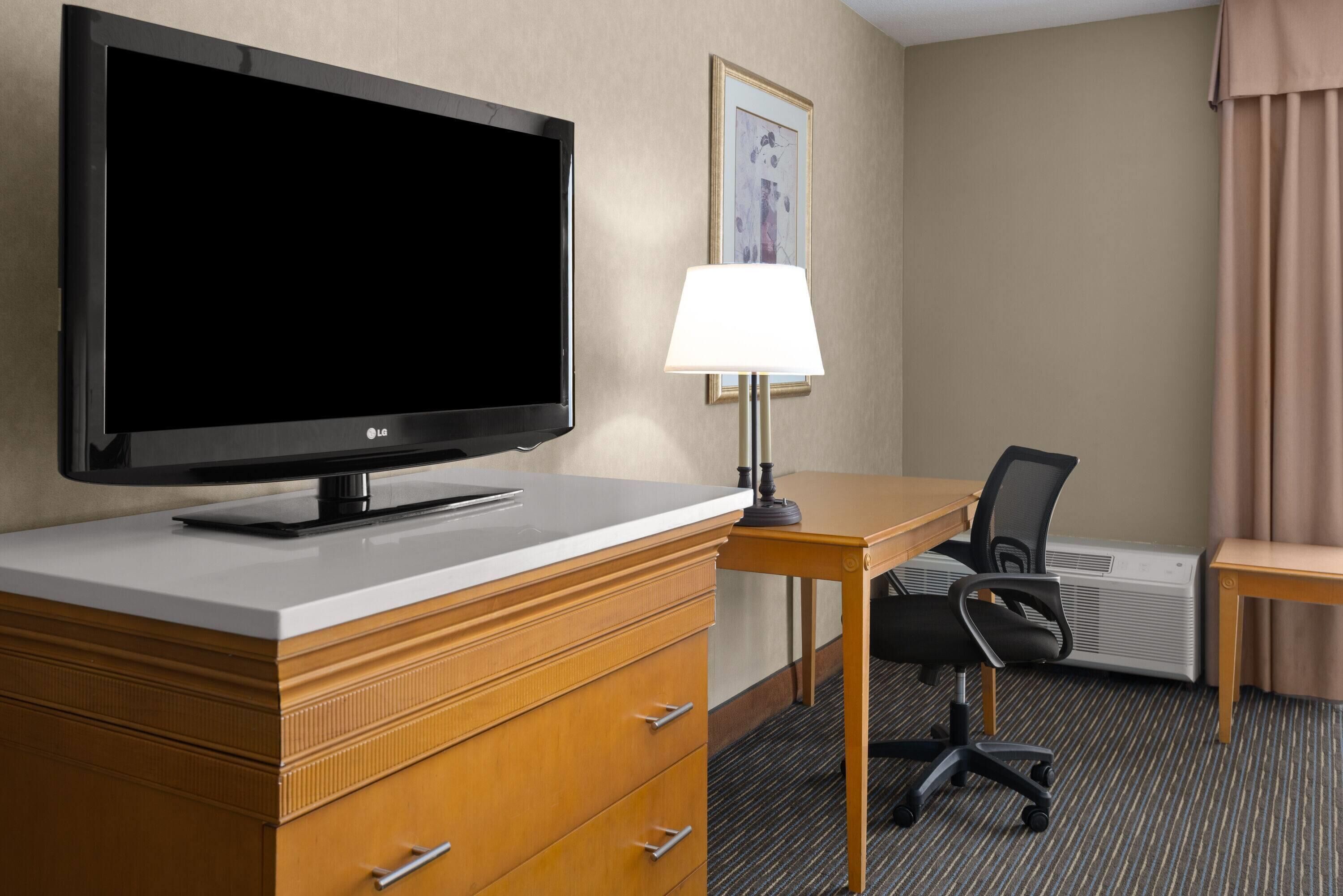 A hotel room with a flat-screen TV, a desk with a chair, a lamp, a framed picture on the wall, and an air conditioning unit.