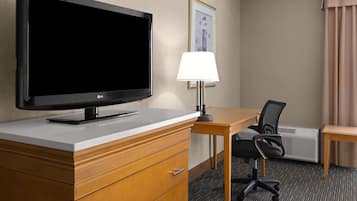 A hotel room with a flat-screen TV, a desk with a chair, a lamp, a framed picture on the wall, and an air conditioning unit.
