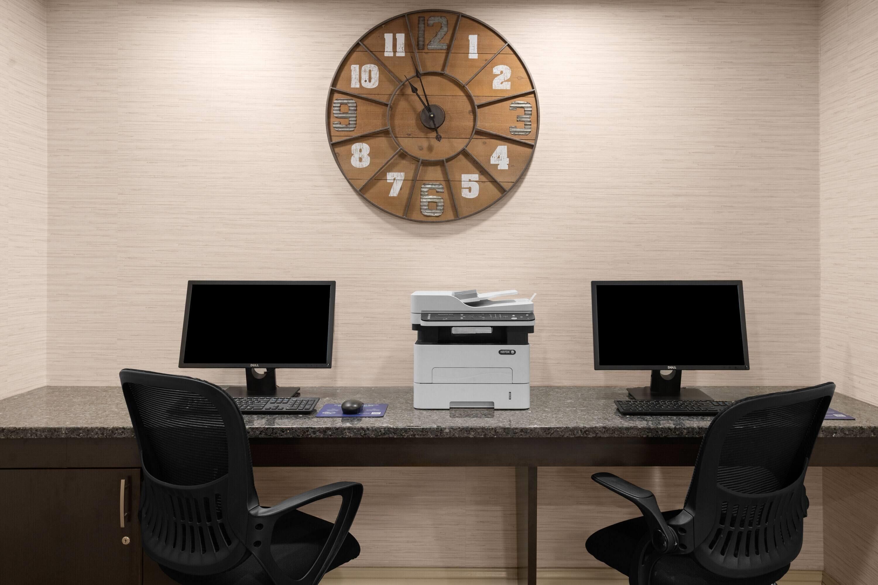 A workspace with two monitors, a printer, and a wooden clock on the wall.