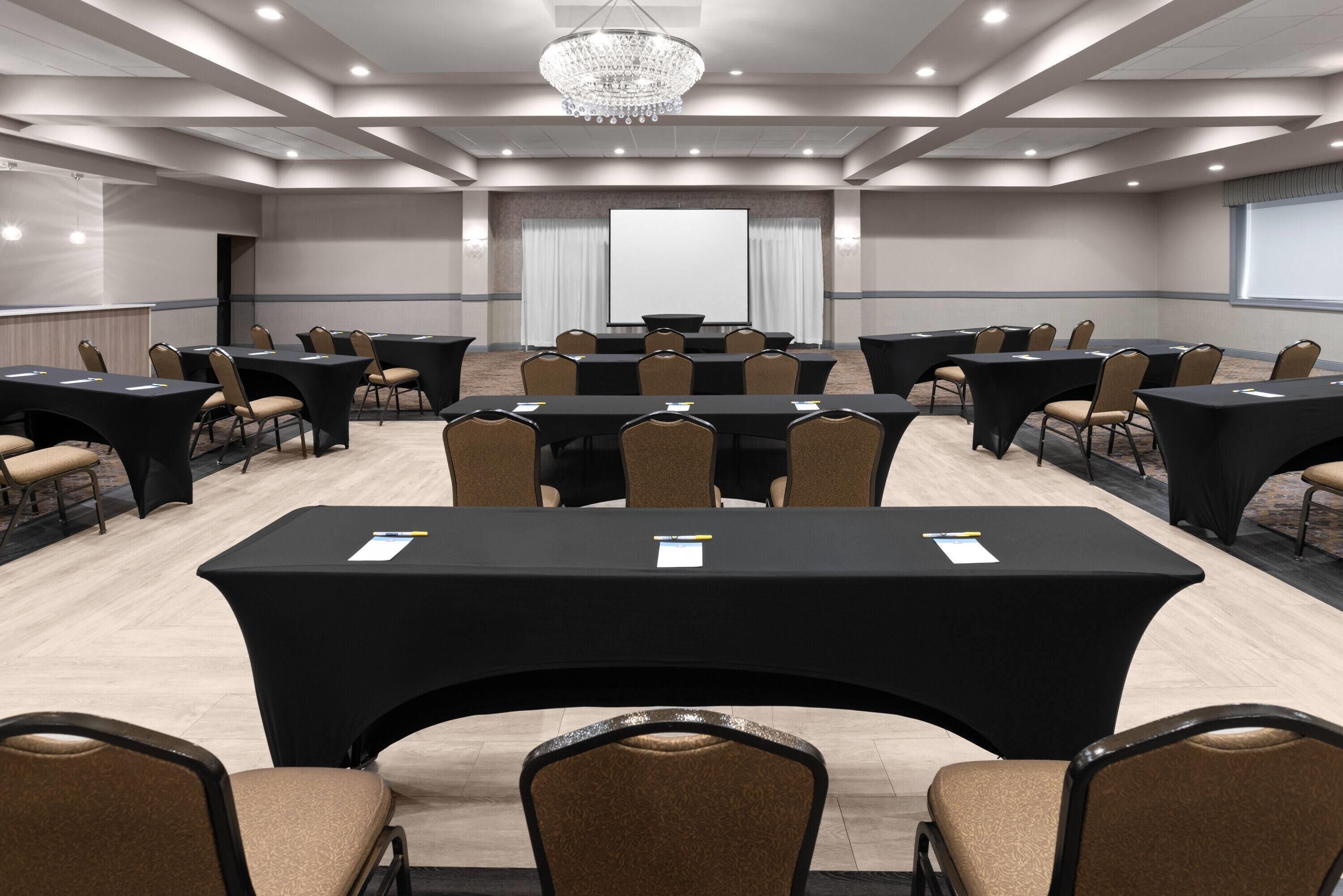 Une salle de conférence équipée de plusieurs tables et chaises, d’un écran de projection et d’un lustre.