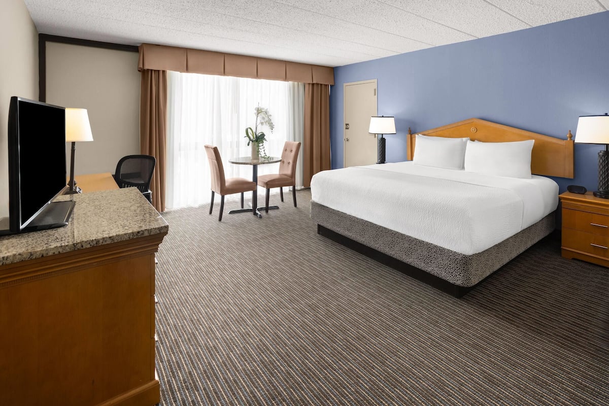 A hotel room with a bed, desk, chair, and a small table with a lamp.