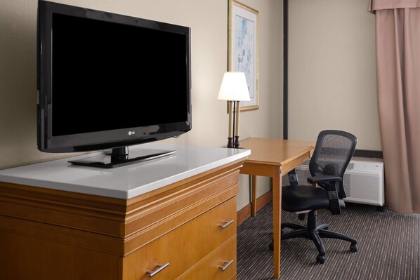 Habitación de hotel con televisión de pantalla plana, cómoda de madera, escritorio, silla, lámpara y un cuadro en la pared.