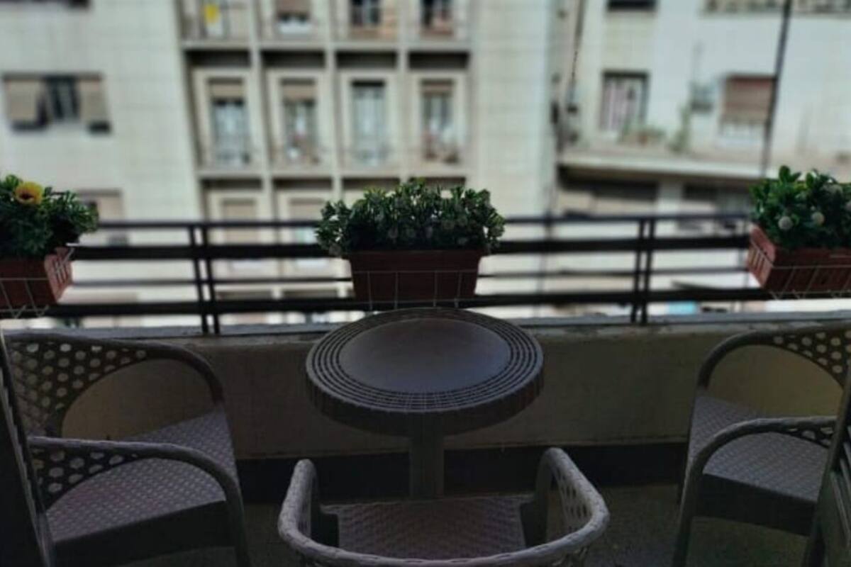 A balcony with two wicker chairs and a small round table, potted plants, and a view of buildings in the background.