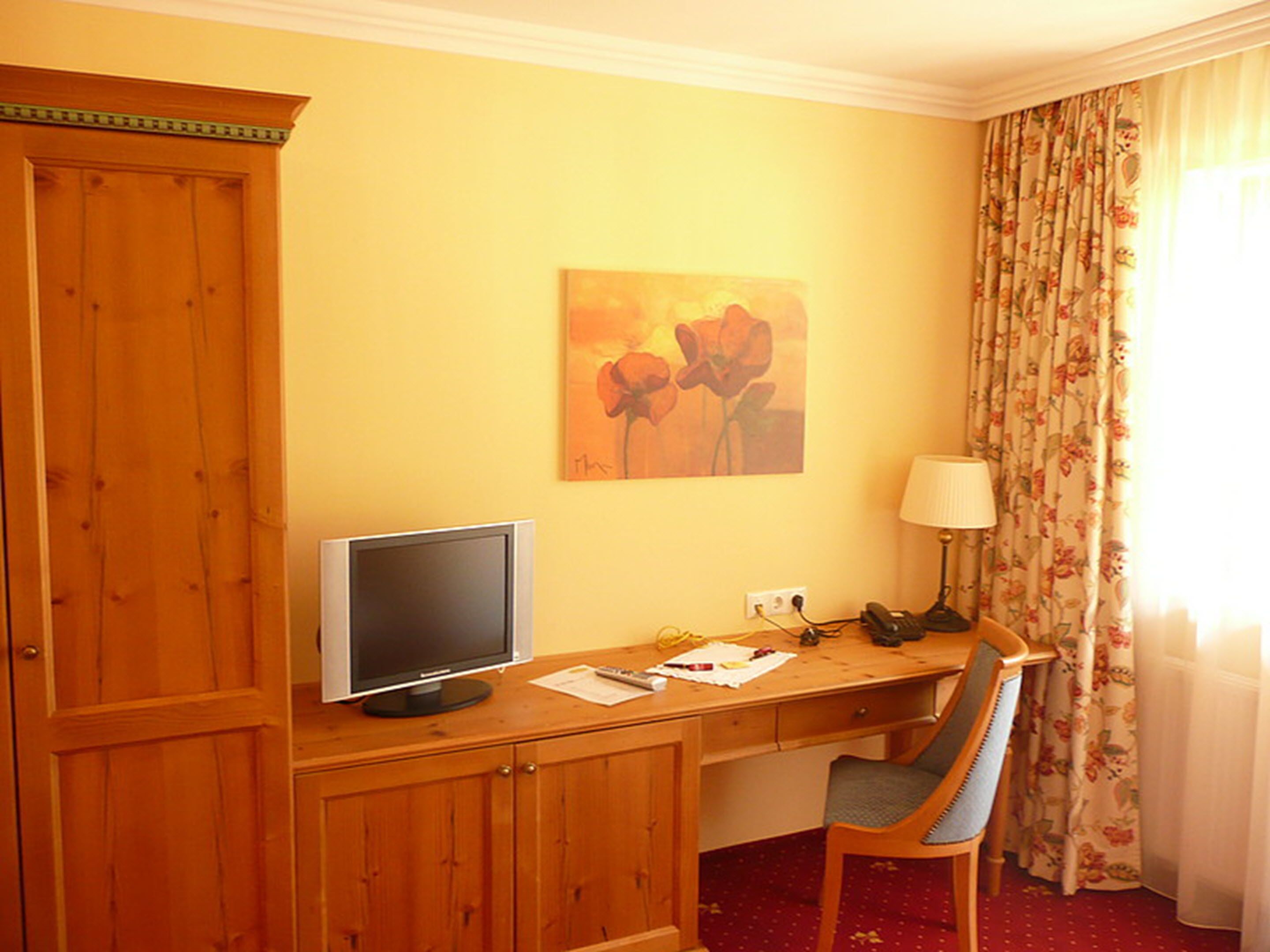 A hotel room with a wooden desk, a television, a chair, a lamp, and a painting on the wall.