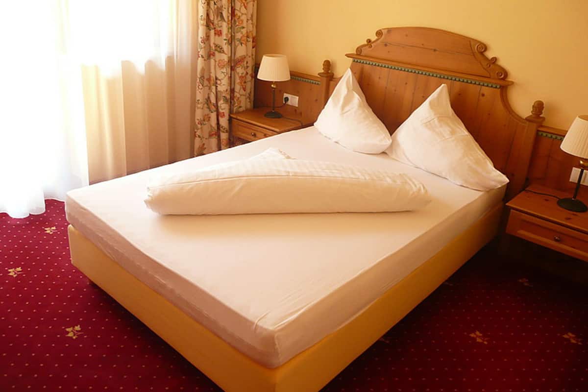 Una cama impecablemente arreglada con sábanas y almohadas blancas, un cabecero de madera y una mesita de noche con una lámpara.