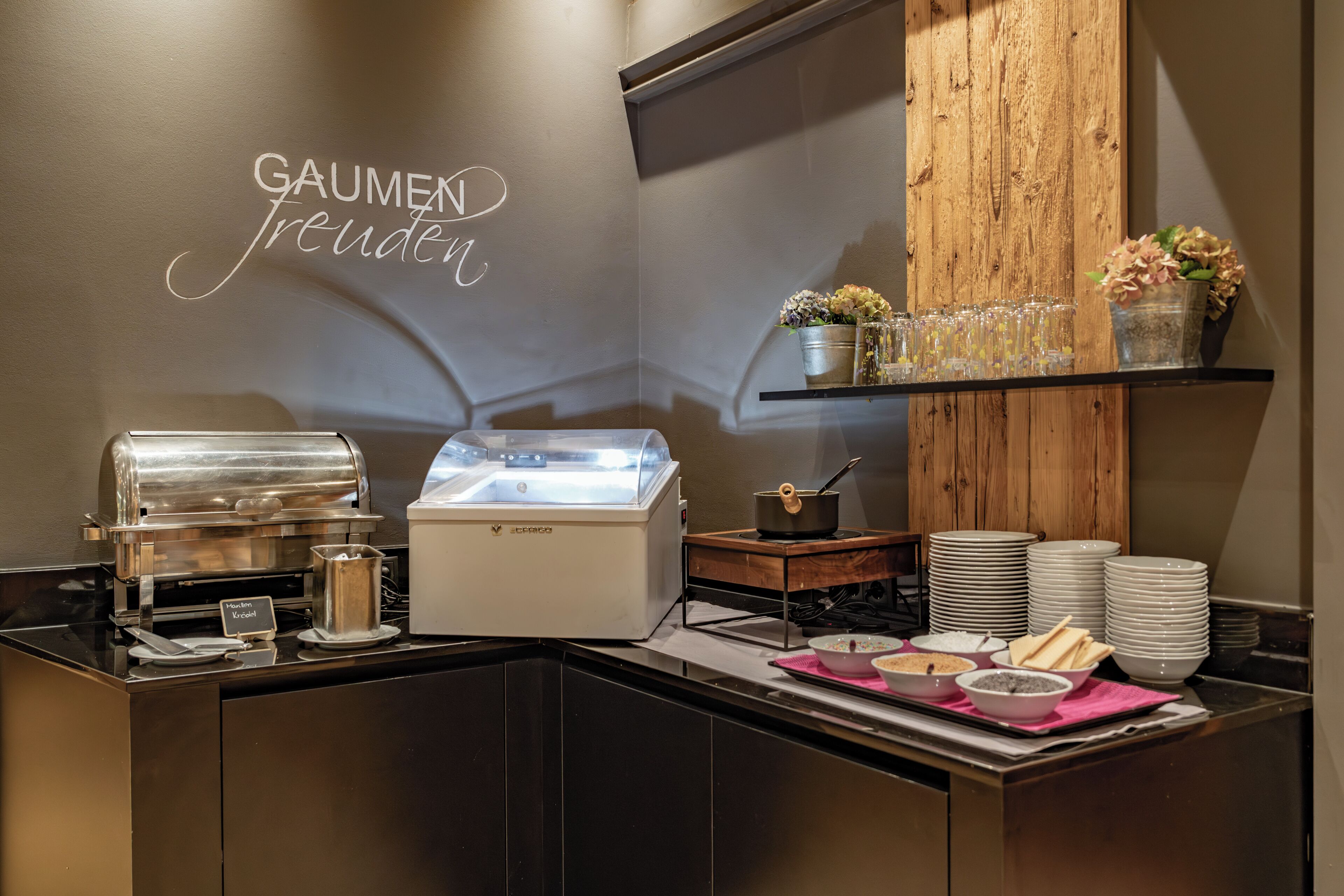 Una barra de desayuno con una estación de café, un cartel que dice 'GAUMEN Freude' y una estantería de madera con flores y vasos.