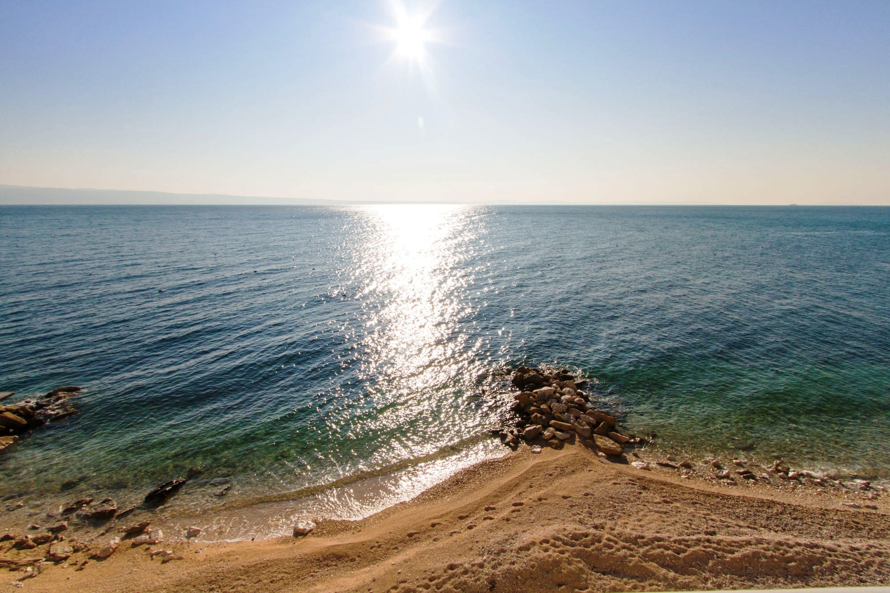 A beach with clear water, a rocky shoreline, and a bright sun in the sky.