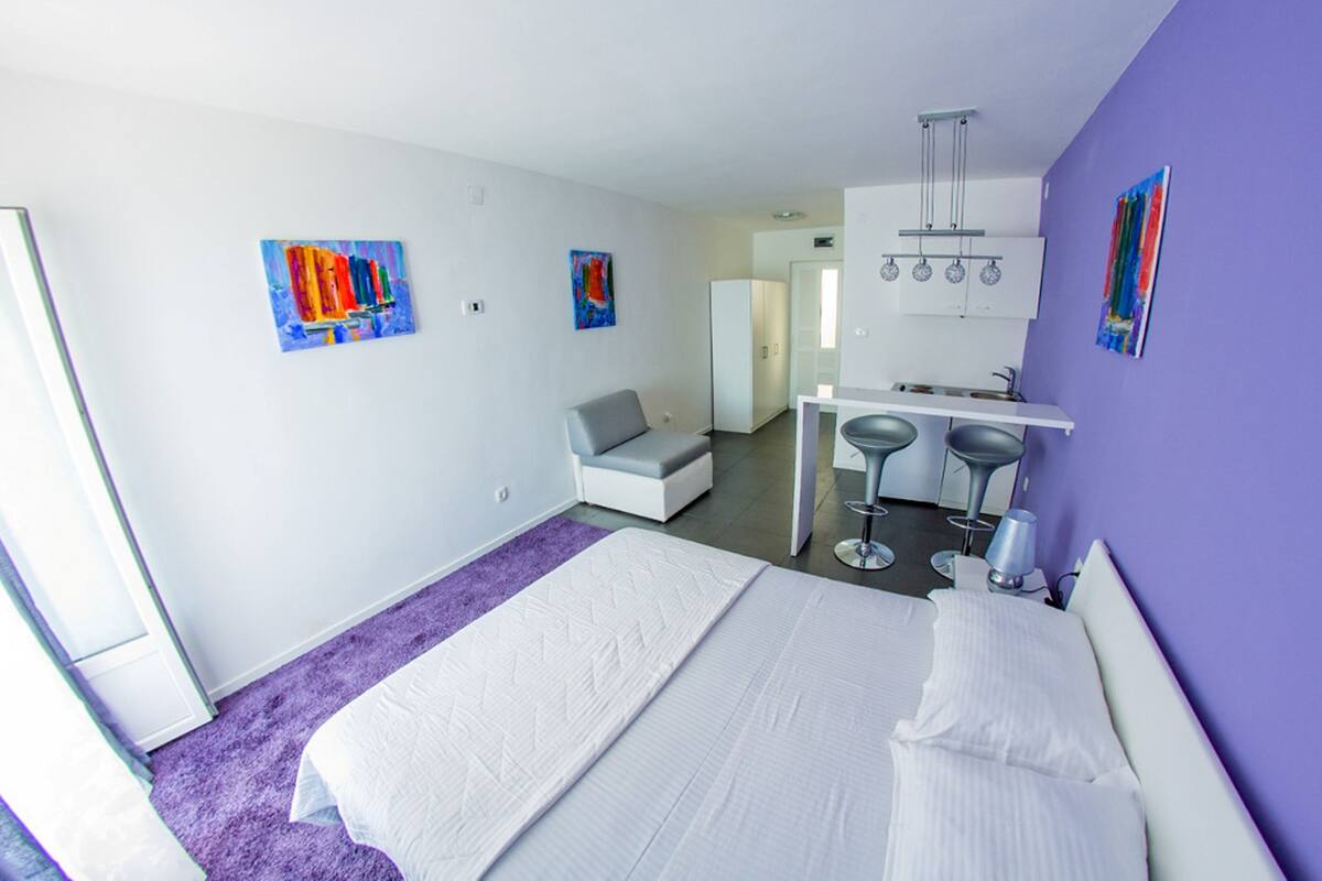 A modern hotel room with a large bed, a small sofa, a kitchenette, and two bar stools.