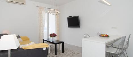 Un salon moderne comprenant un canapé, une table à manger avec des chaises, un téléviseur fixé au mur et une petite table sur laquelle est posé un vase de fleurs.