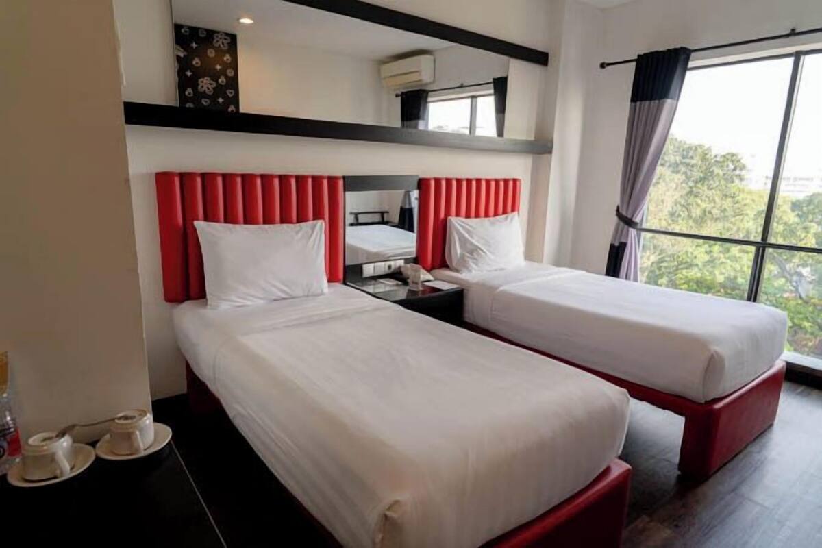 A hotel room with two beds, a red headboard, a nightstand, and a window with curtains.