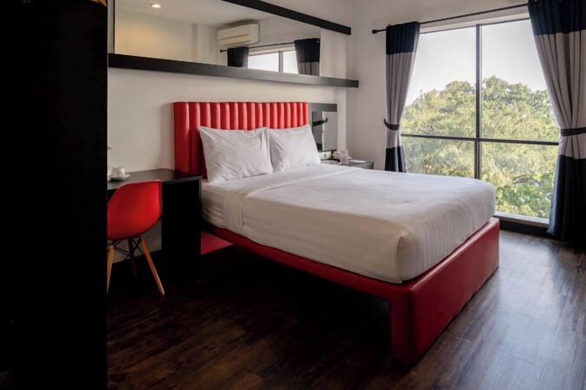 A modern hotel room with a red bed frame, a desk with a red chair, and a large window offering a view of greenery.