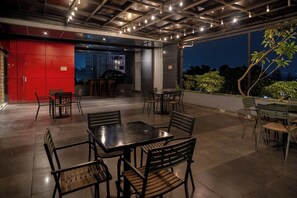 An outdoor dining area with tables and chairs, a red wall, and a view of the city at night. - Kalya Hotel Bandung (Bandung)