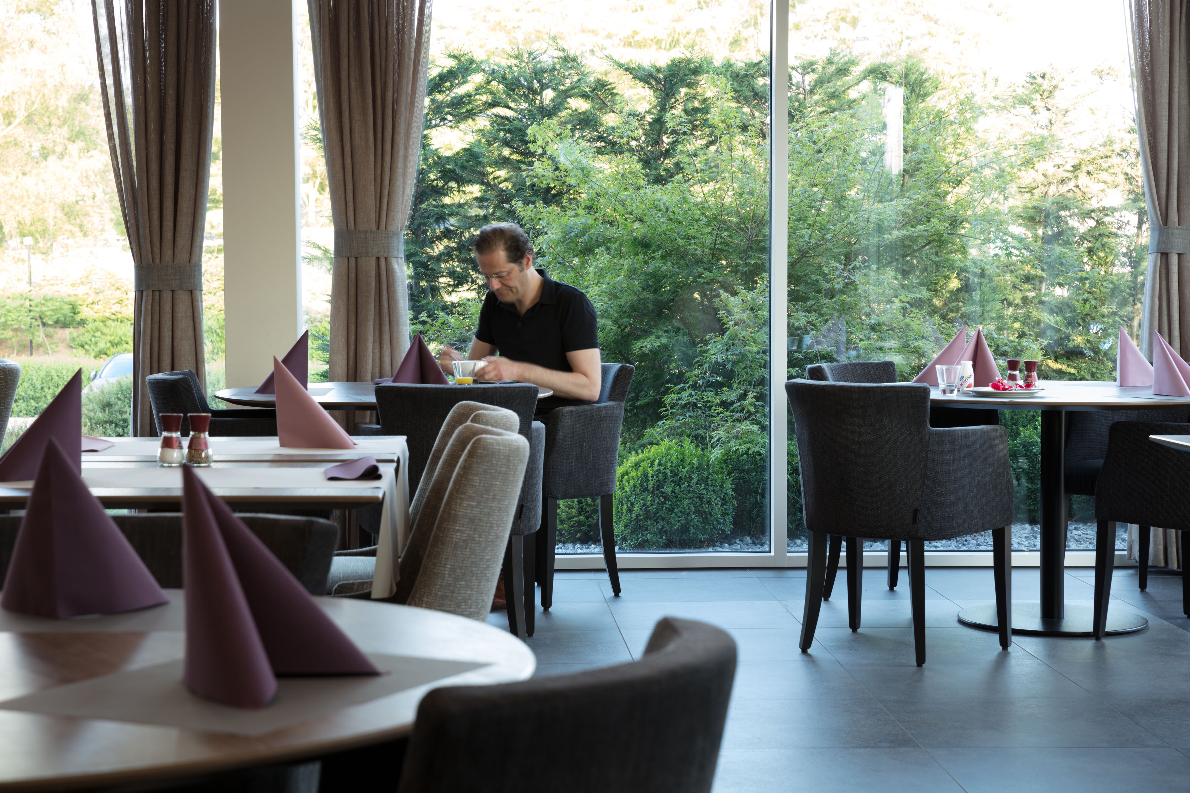 A restaurant with tables set for dining, featuring a view of greenery through large windows.