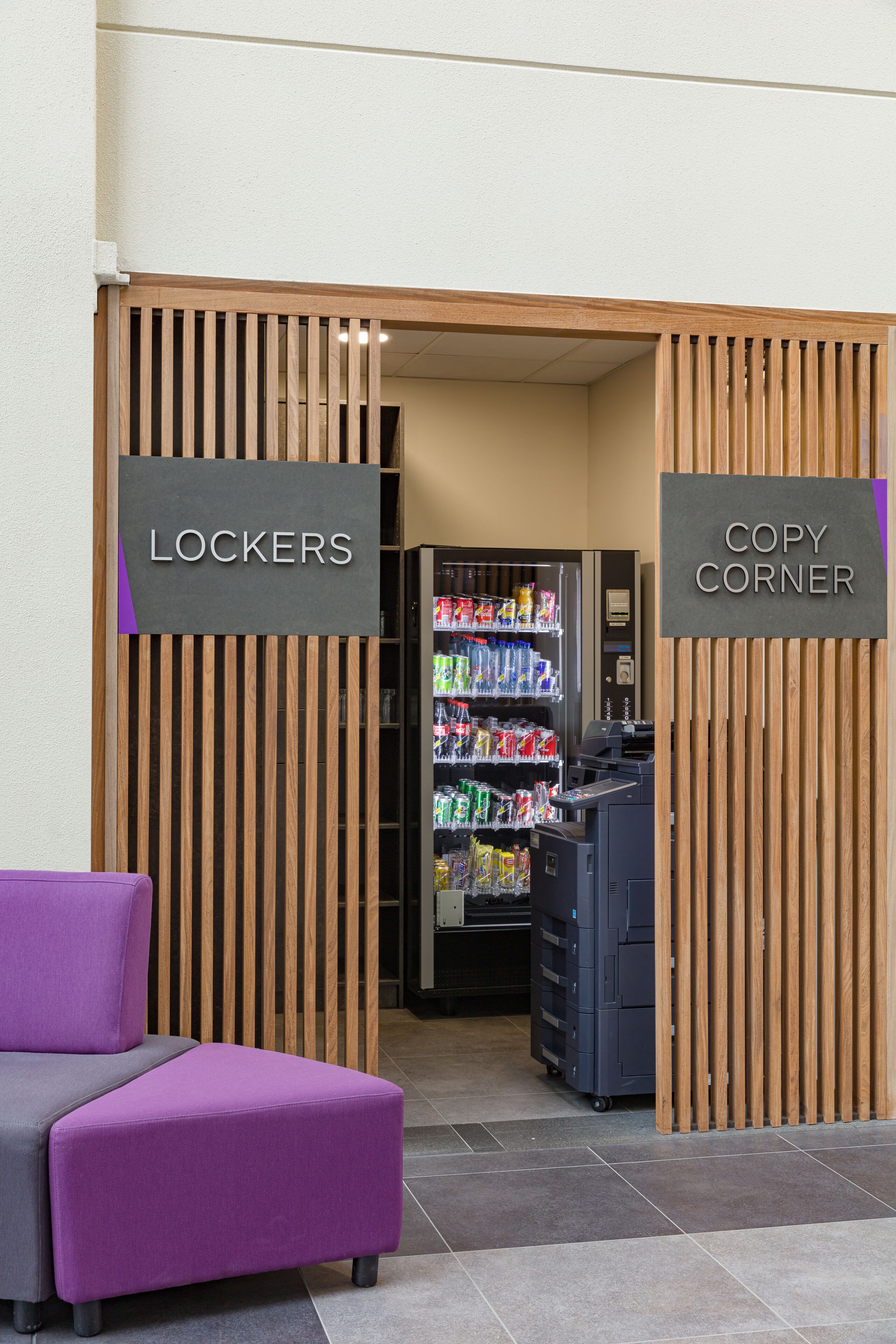 Un hall d’hôtel avec un distributeur automatique, un espace détente aux couleurs violettes et des panneaux indiquant les « CASIERS » et le « SERVICE DE PHOTOCOPIE ».