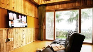 A room with a wooden interior, a leather sofa, a television on the wall, and a view of palm trees through large windows. - Memory In Jeju Pension (Seogwipo)