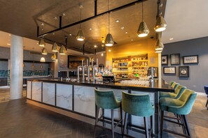 Een bar met een marmeren toonbank, groene barstoelen, hanglampen en een bar gevuld met flessen.