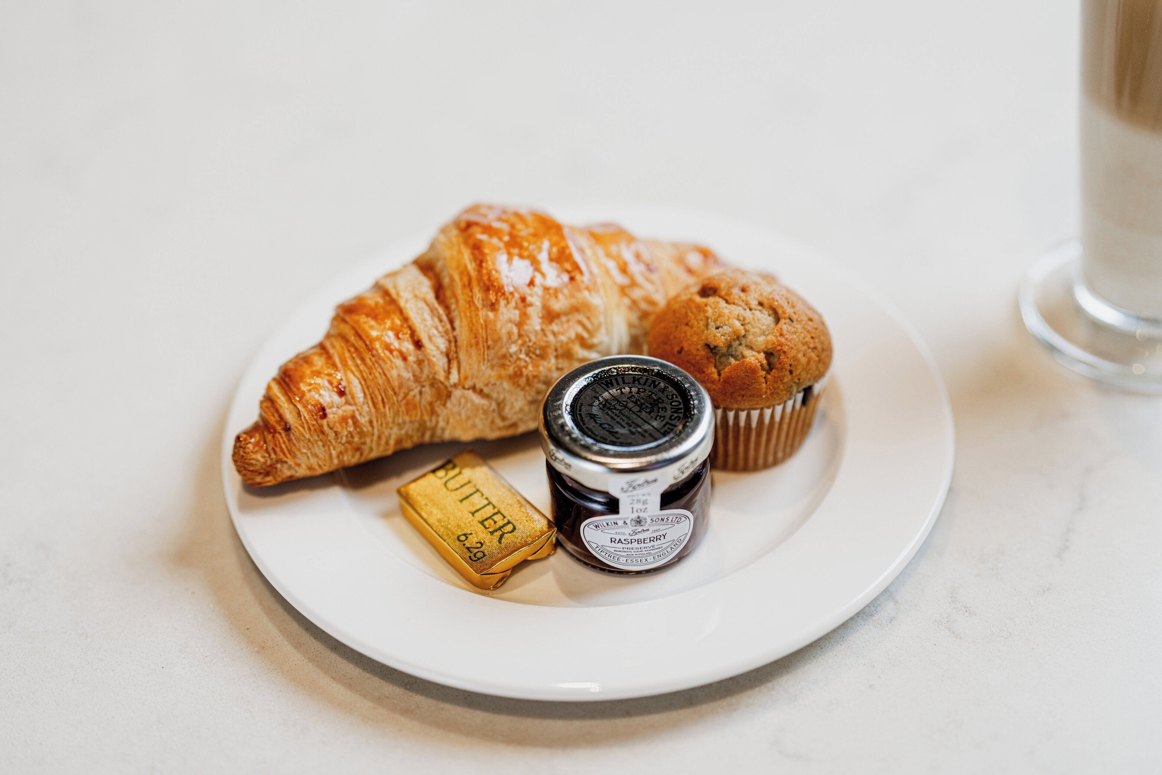 Un plato con un croissant, un muffin y un frasco de mermelada de frambuesa.