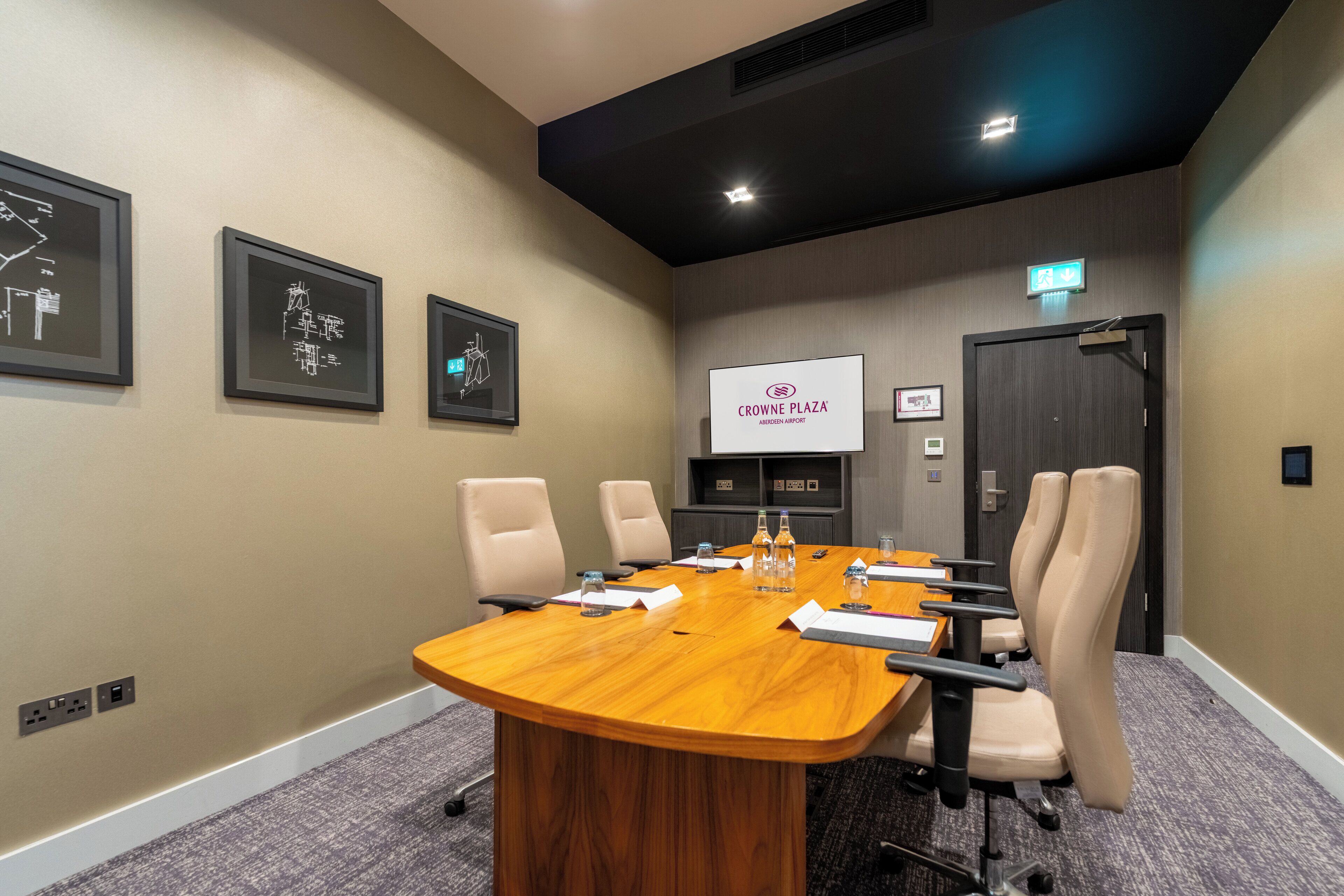 A conference room with a long wooden table, beige chairs, and a projector screen displaying "CROWNE PLAZA AMBLEDEN AIRPORT".