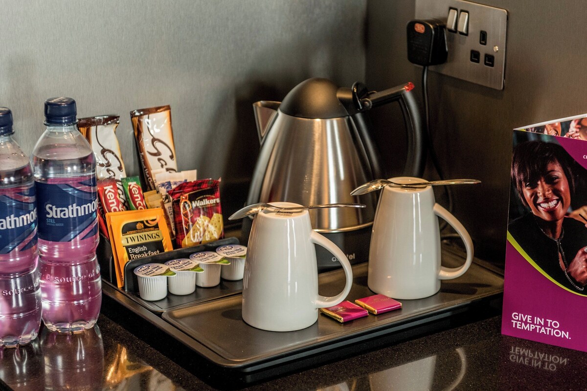 Ontbijt in een hotelkamer met waterflesjes, snacks, een koffiezetapparaat en een reclamebord.