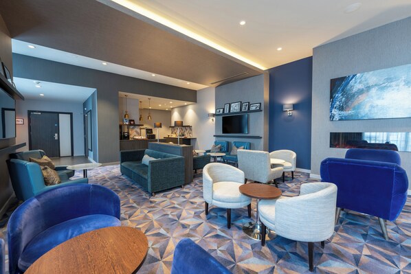 A modern hotel lobby with a geometric patterned floor, various seating options including sofas and armchairs, a television mounted on the wall, and a kitchen area visible through an open door. - Crowne Plaza Aberdeen Airport by IHG (Aberdeen)