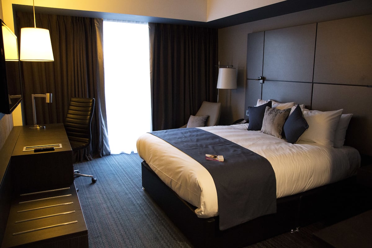Une chambre d’hôtel avec un lit, un bureau, une chaise, une lampe et une fenêtre avec des rideaux.