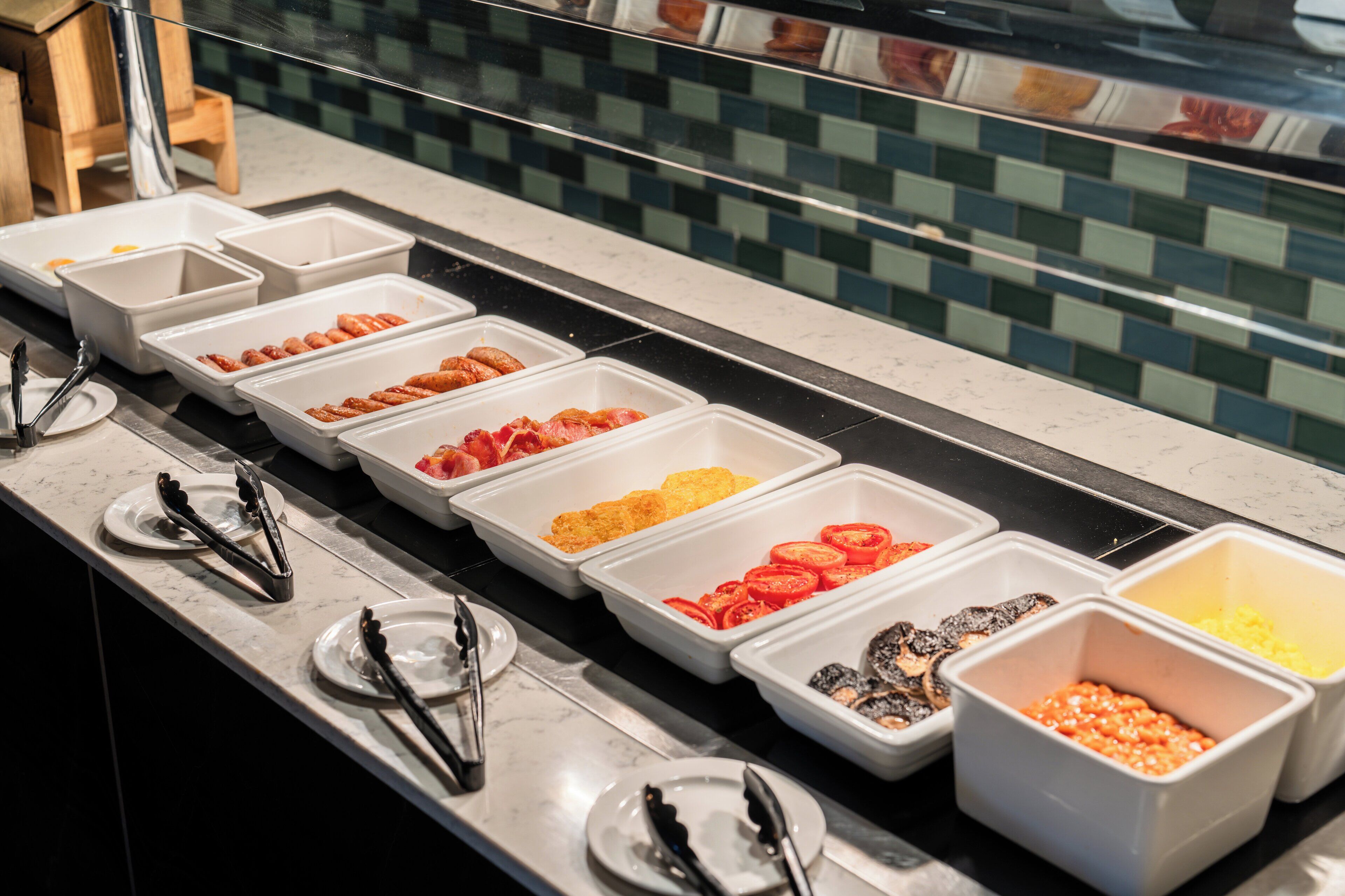 A buffet line with various food items in white trays, including sliced tomatoes, mushrooms, and other vegetables.