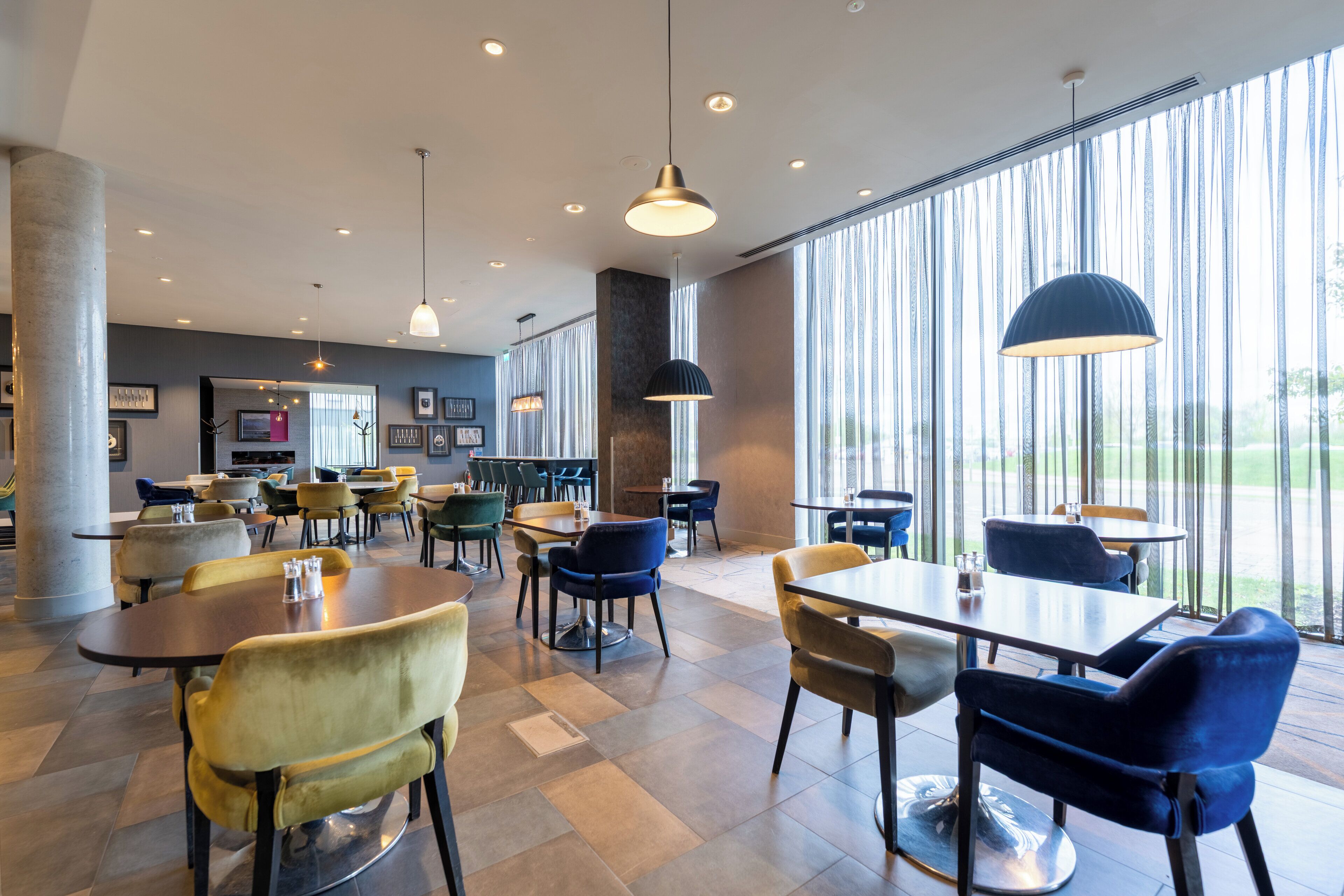 A modern dining area with multiple tables and chairs, large windows with vertical blinds, and pendant lighting.