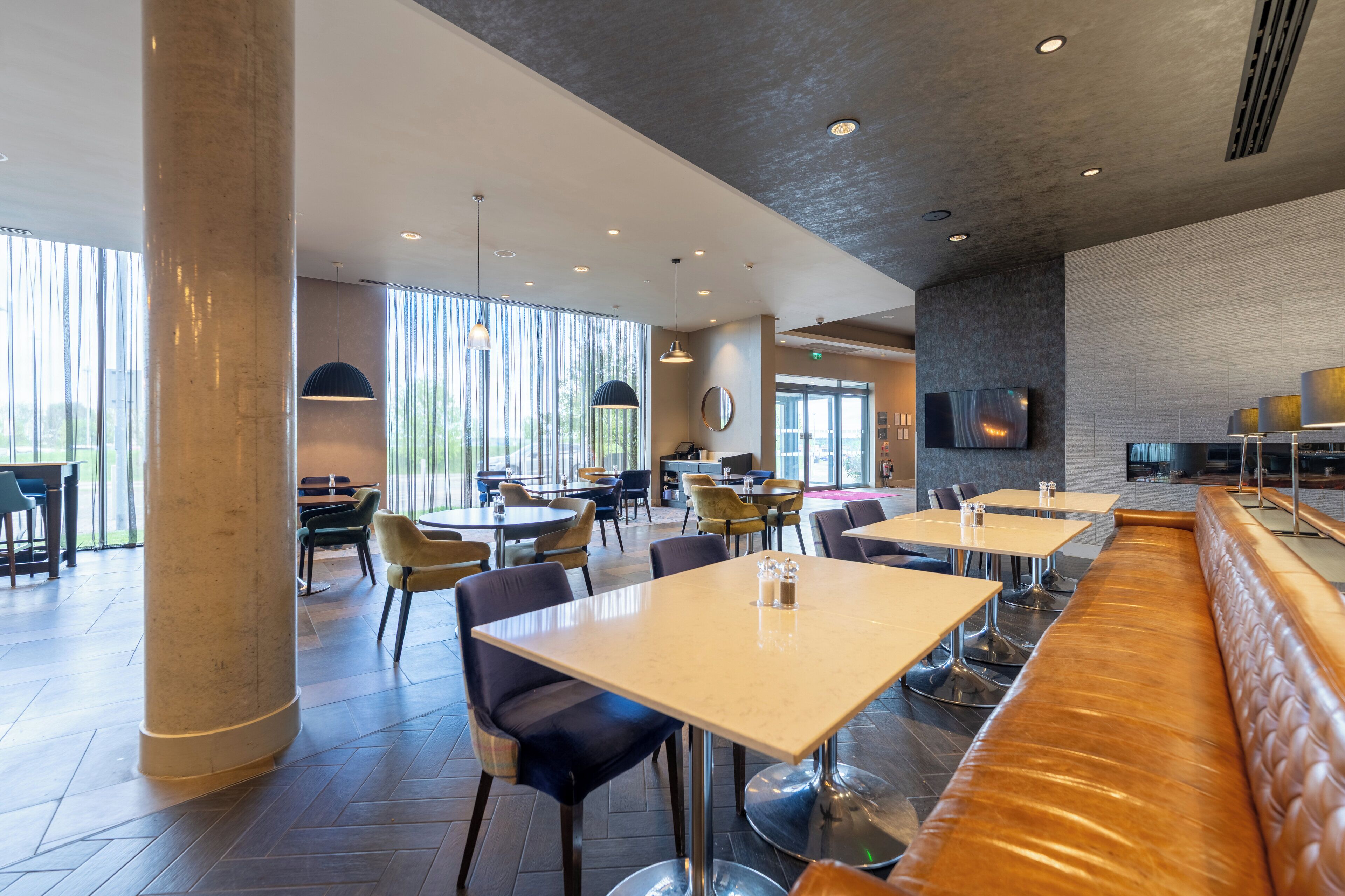 Ein modernes Restaurant mit hohen Decken, großen Fenstern und einer Auswahl an Sitzgelegenheiten, darunter Tische und eine lange Bank.