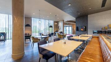 Ein modernes Restaurant mit hohen Decken, großen Fenstern und einer Auswahl an Sitzgelegenheiten, darunter Tische und eine lange Bank.