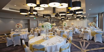 Un salón de banquetes con mesas redondas, manteles blancos, cintas doradas y centros de mesa florales.