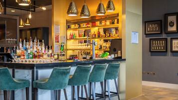 Zona de bar con mostrador, taburetes verdes, lámparas colgantes y estanterías llenas de botellas y vasos.