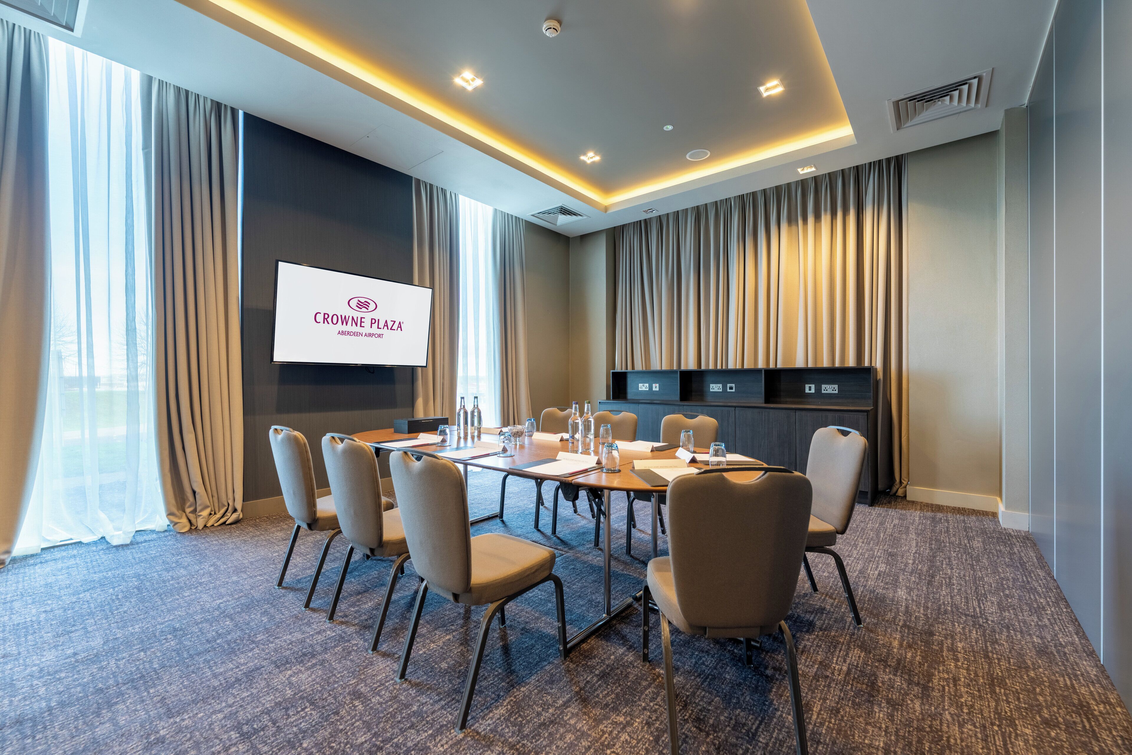 A conference room with a large screen displaying "CROWNE PLAZA," beige chairs, a long table, and a carpeted floor.