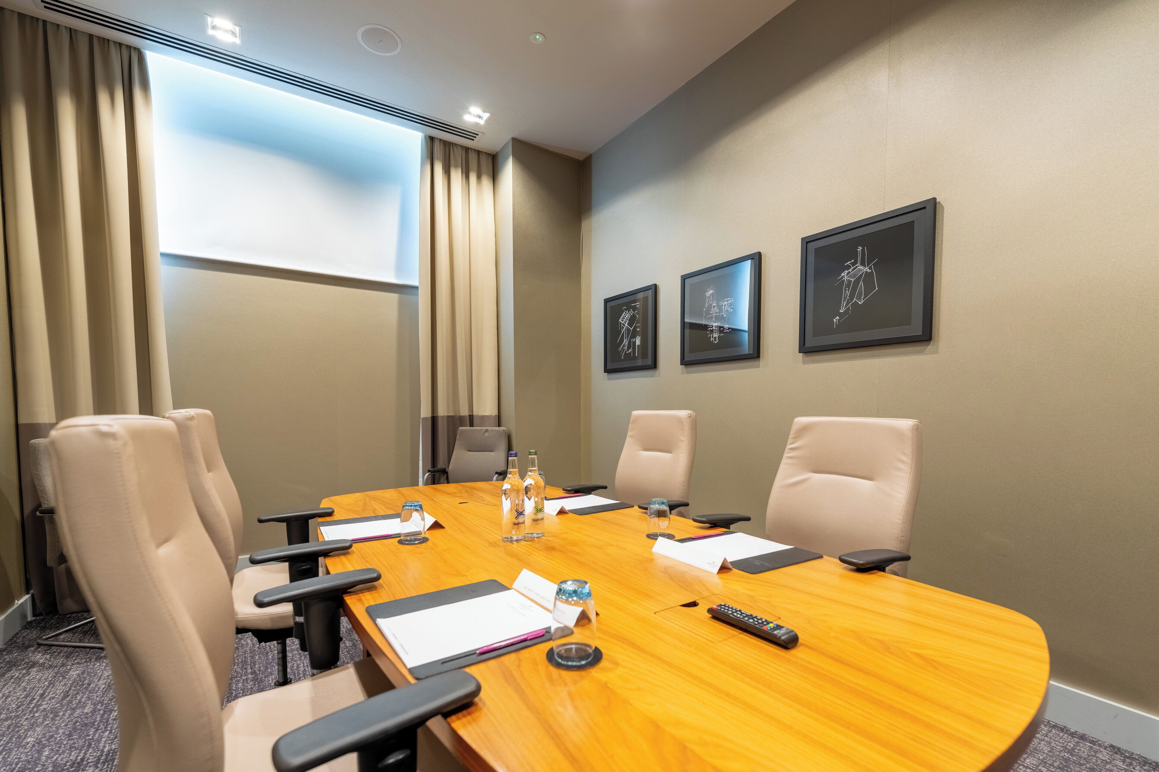 A conference room with a wooden table, beige chairs, framed artwork on the wall, and a window with curtains.
