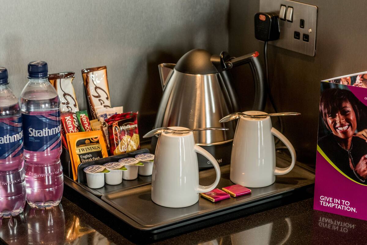 Une table de petit-déjeuner installée dans une chambre d'hôtel, avec des bouteilles d'eau, des en-cas, des cafetières et un panneau publicitaire.