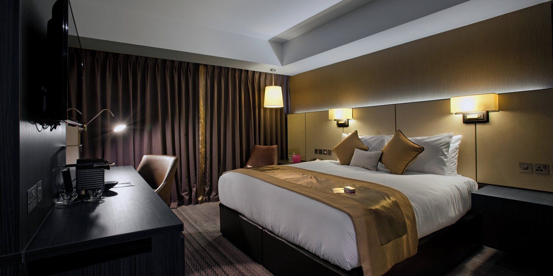 A hotel room with a bed, bedside lamps, a desk, and a television.
