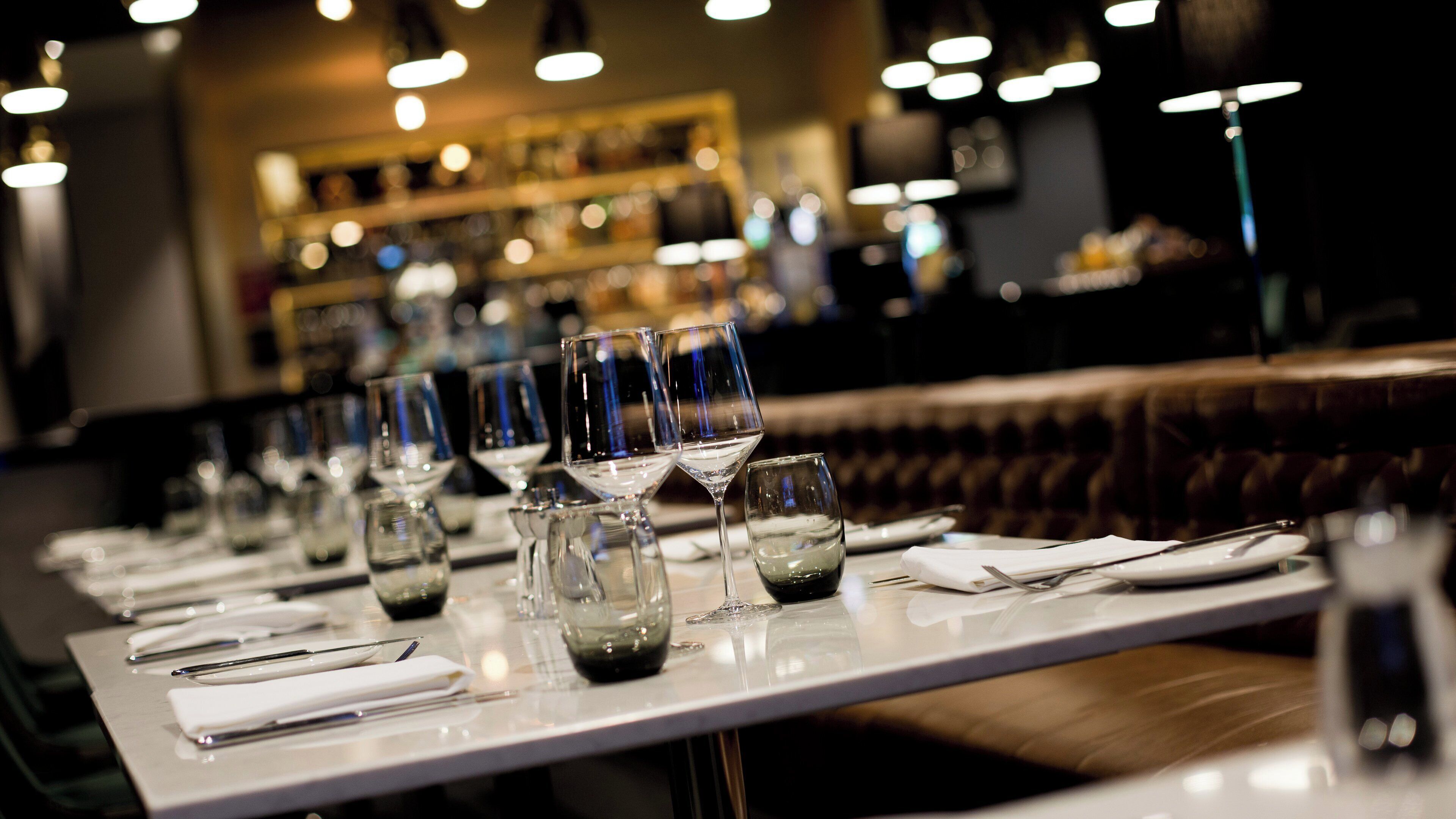 L’intérieur d’un restaurant, avec une longue table dressée pour le dîner, agrémentée de nombreux verres à vin et de verres à eau.
