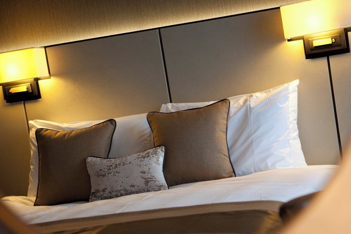 A hotel room with a bed, pillows, and two wall-mounted lamps.