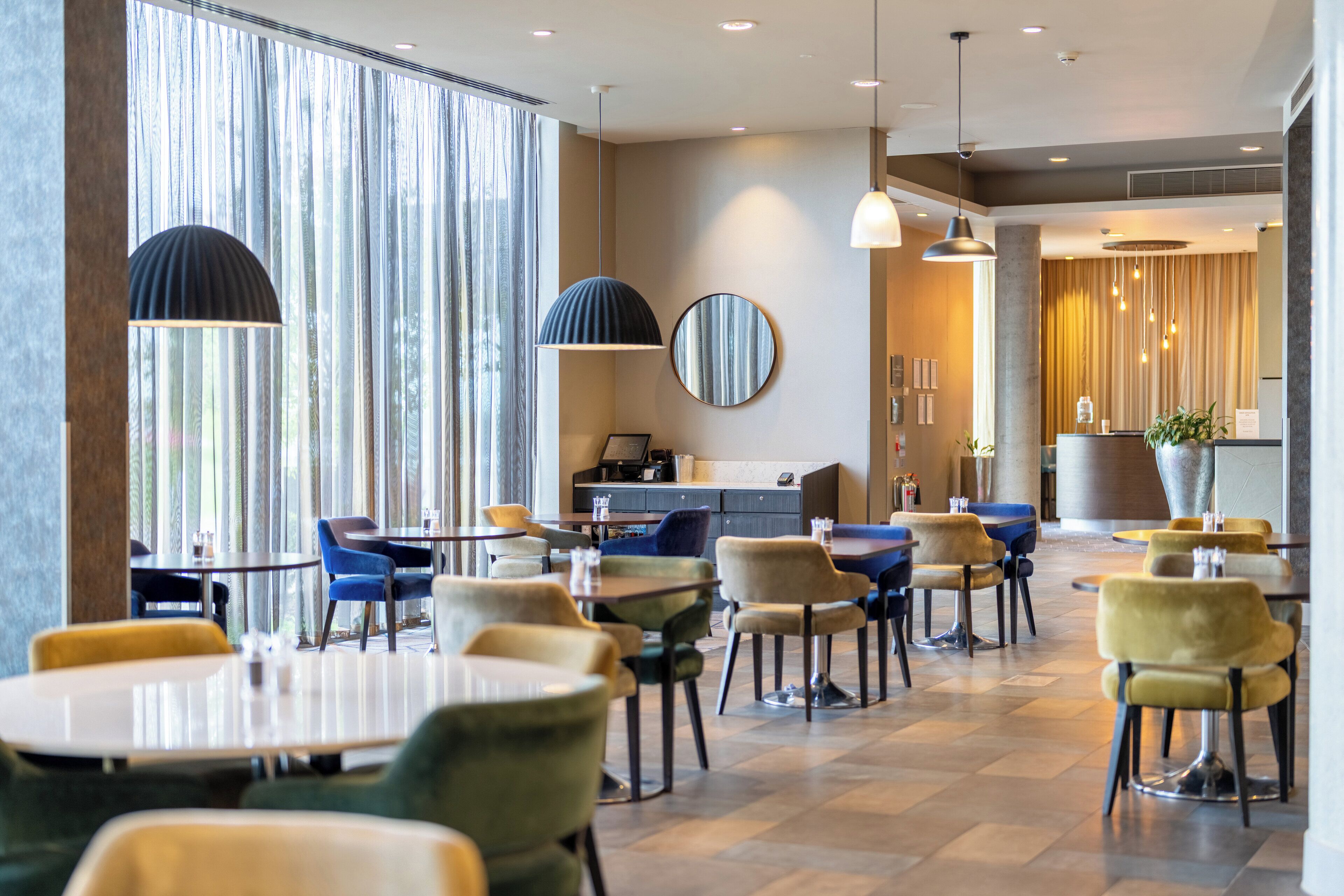 A modern hotel lobby with round tables, comfortable chairs, and large windows with curtains.