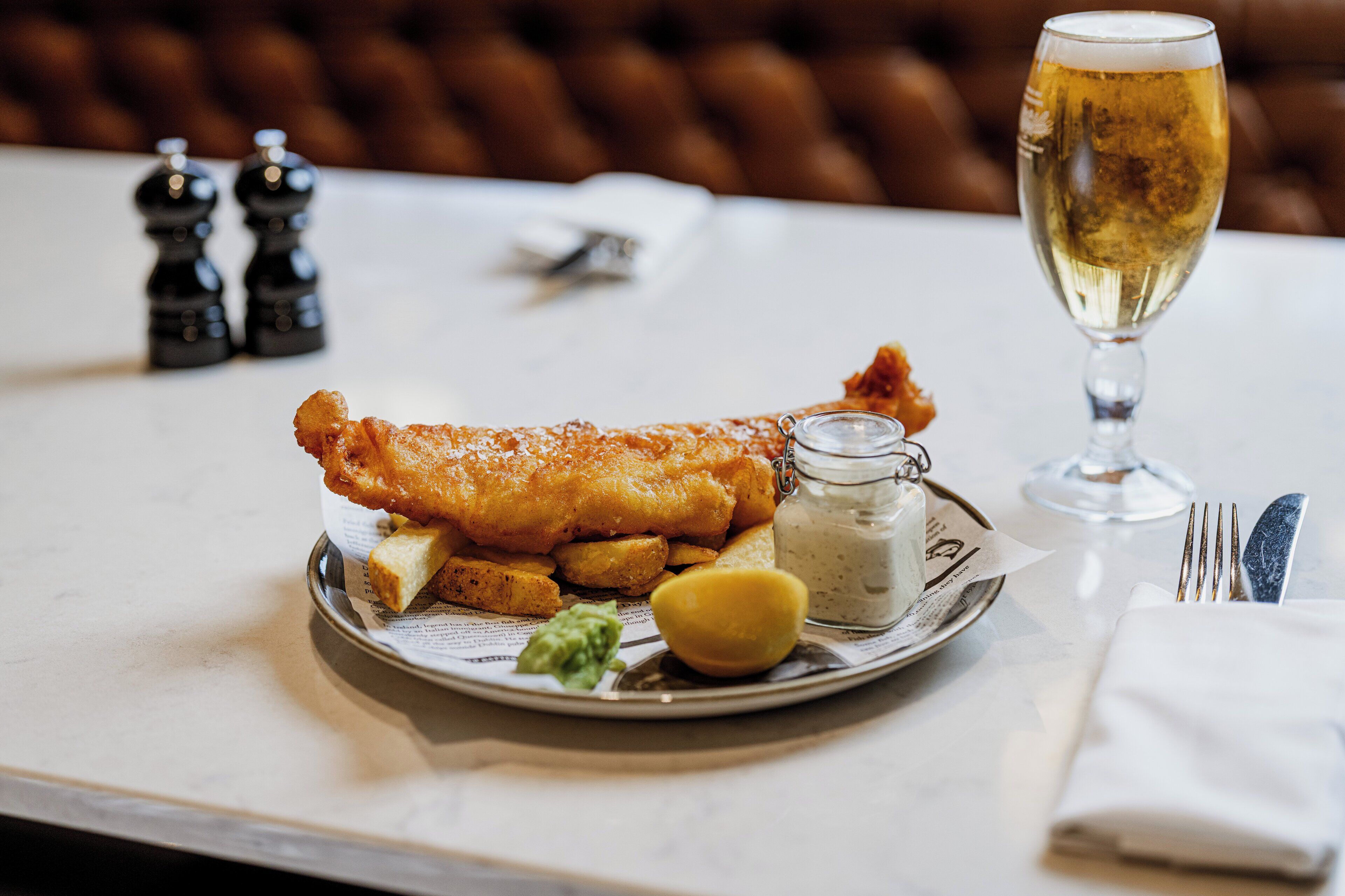 A plate of fish and chips with a glass of beer and condiments on a table.