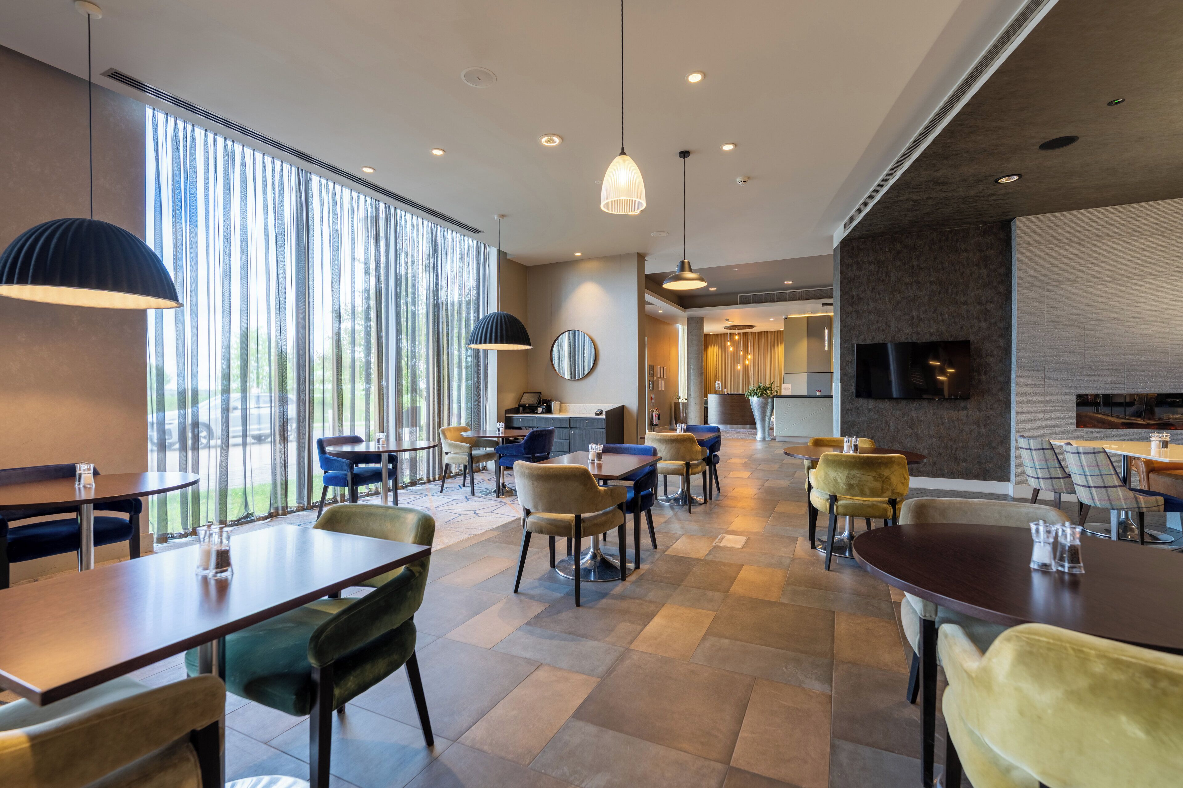 A modern dining area with multiple tables and chairs, large windows, and a television on the wall.