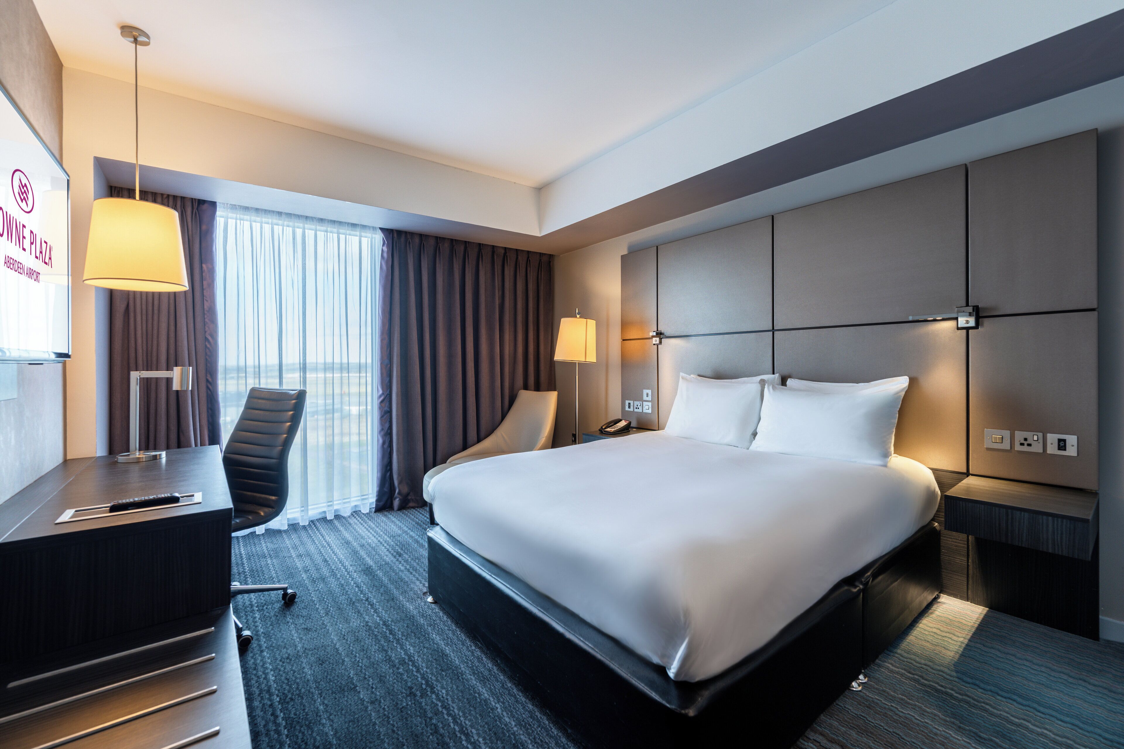 A modern hotel room with a large bed, a desk, a chair, and a large window with curtains.