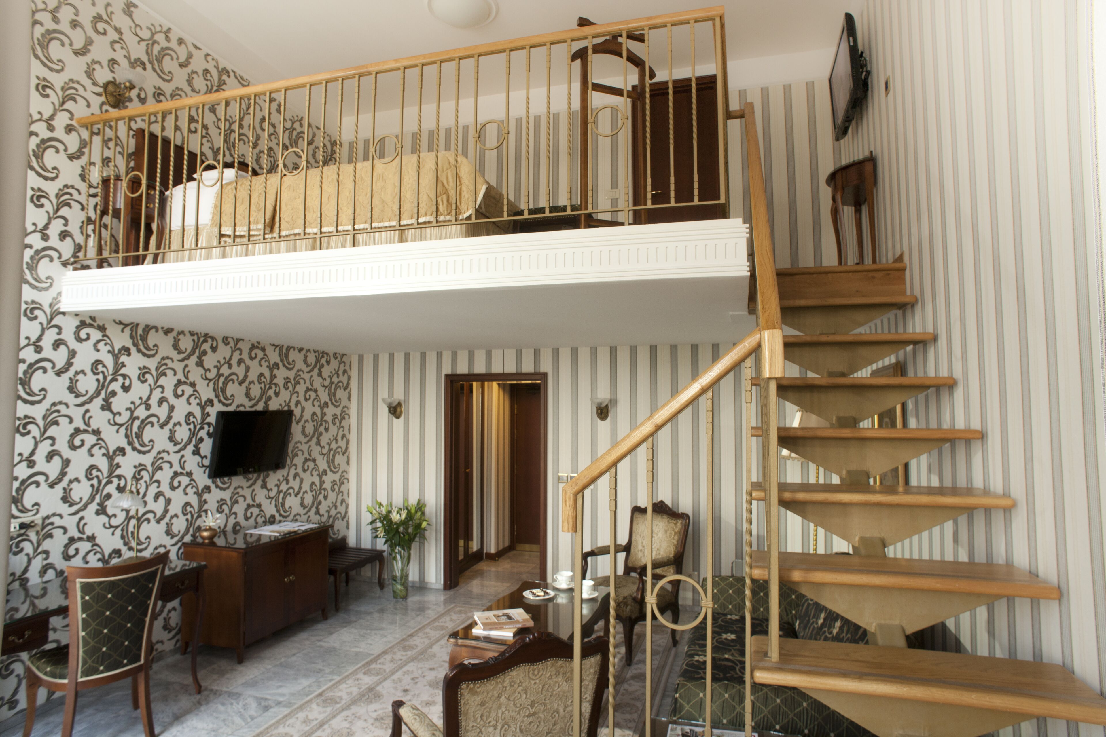 A spacious room with a high ceiling, a wooden staircase, a flat-screen TV mounted on the wall, and a patterned wallpaper design.