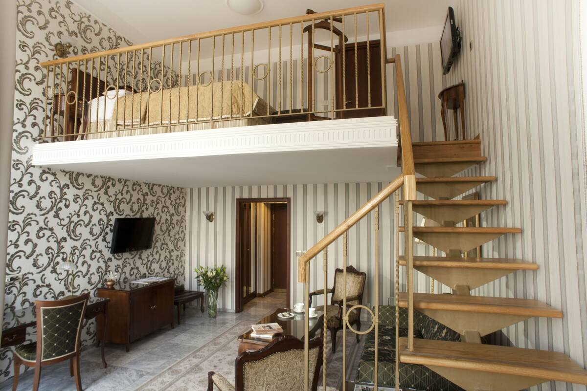 A spacious room with a high ceiling, a wooden staircase, a balcony, and a television mounted on the wall.